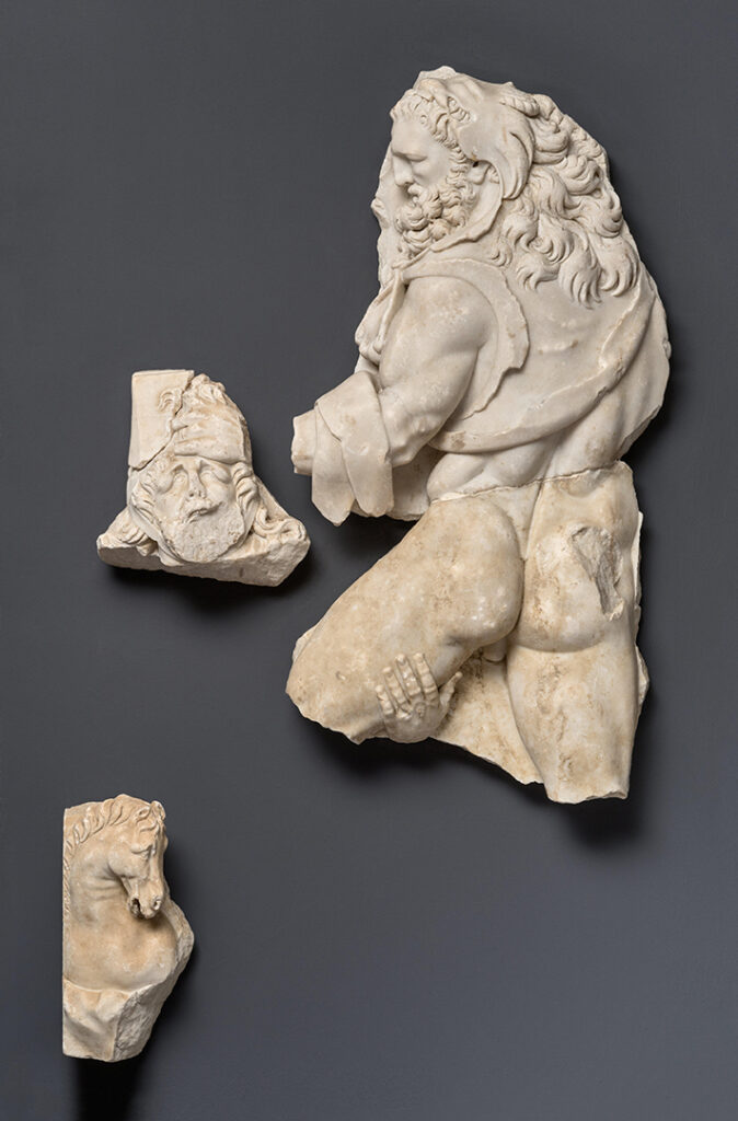 Relief représentant Hercule et le roi Diomède. Le relief est en marbre blanc. Il est en forme de rectangle, à la verticale, et la bordure est sculptée comme un cadre. Plusieurs parties du relief ont été cassées et sont manquantes. Hercule est représenté de dos. Sa tête et une partie de son dos sont couverts par la peau du lion de Némée. La crinière du lion prolonge la chevelure d’Hercule. Hercule empoigne par les cheveux le roi Diomède, qui s’accroche à la cuisse nue d’Hercule. En bas à gauche, la tête d’une jument est représentée.