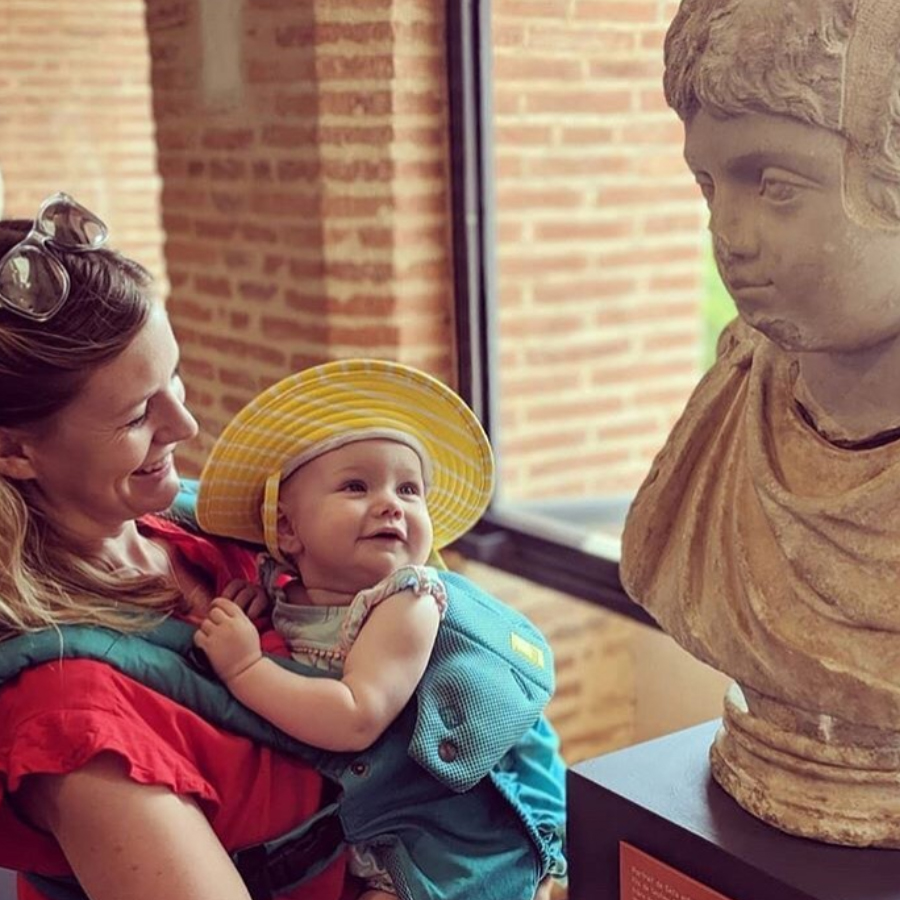 Un bébé, dans les bras d'une femme, souris en regardant un buste romain sculpté.