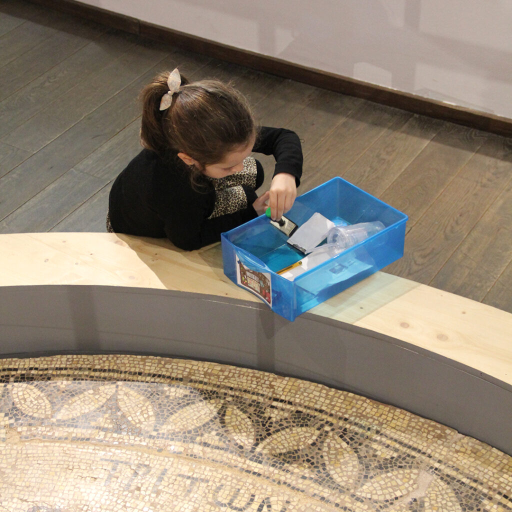 Une petite fille saisit un tampon encreur dans une boîte en plastique. Elle est appuyée sur le rebord de l'encadrement de la mosaïque de Saint-Rustice.