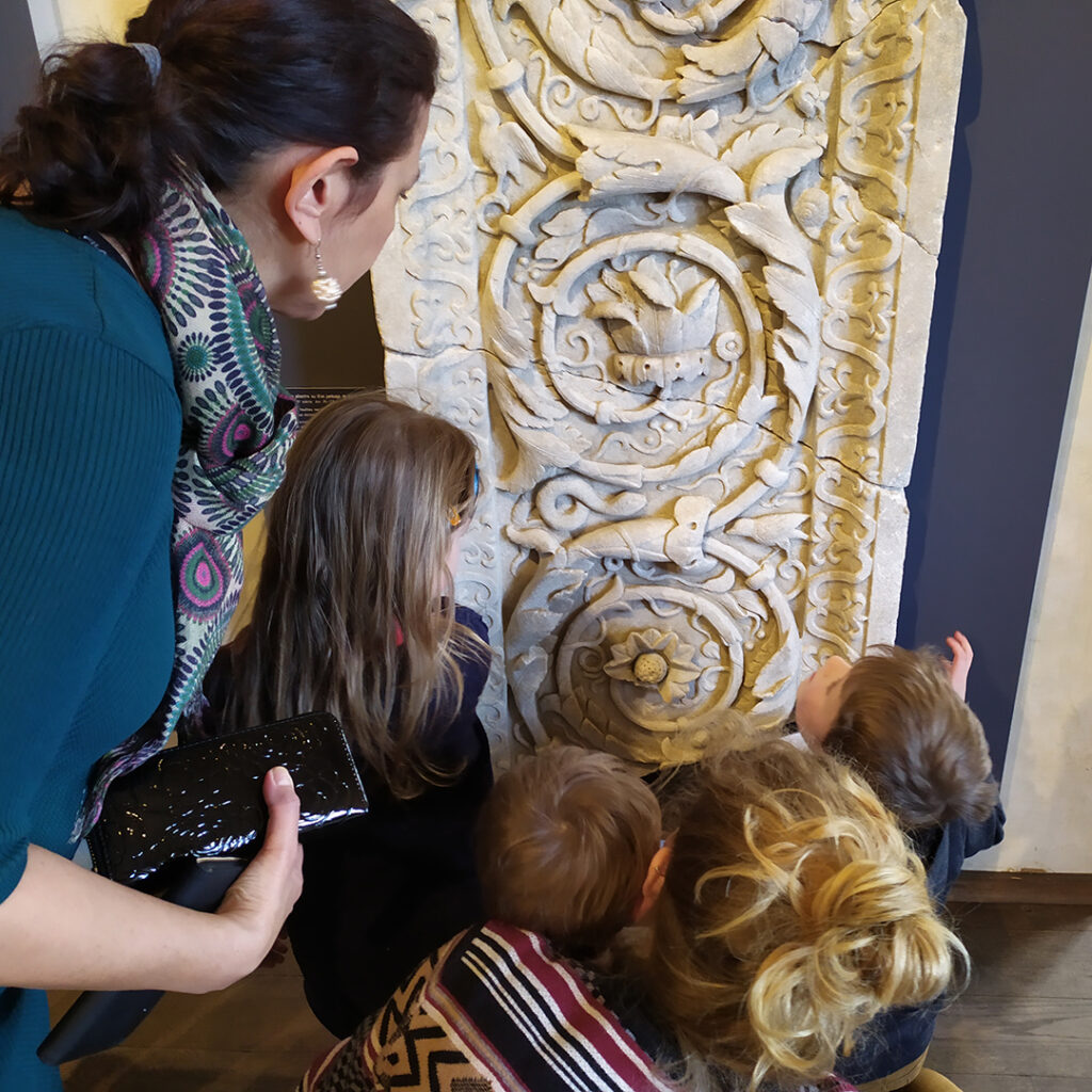 Trois jeunes enfants, de dos, et une femme adulte, cherchent des animaux représentés sur un pilastre sculpté en marbre.