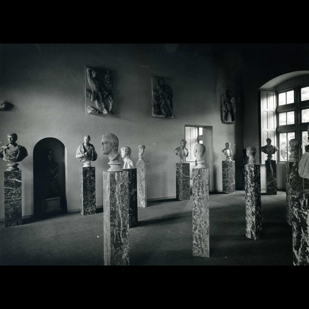 Photographie d'une ancienne présentation du musée Saint-Raymond, dans les années 1950. On voit des portraits romains sur des colonnes en marbre et des reliefs des travaux d'Hercule accrochés en hauteur sur un mur.