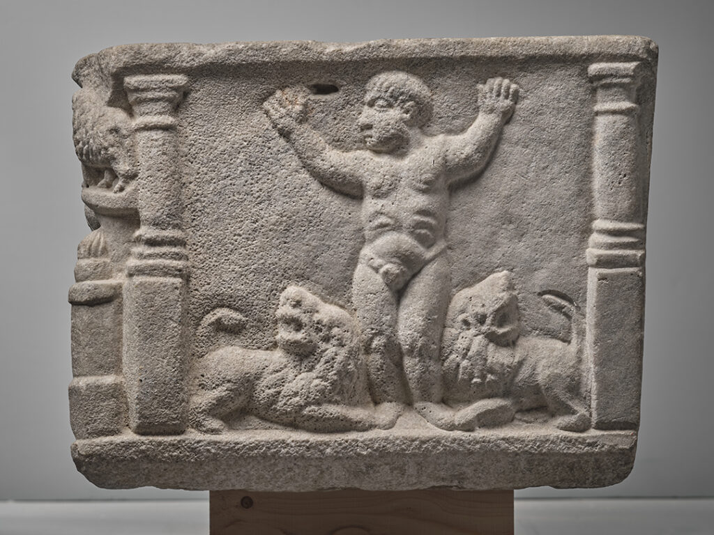 Face latérale droite de la cuve d'un sarcophage. Au centre, un homme nu vue de face, Daniel, lève les bras en l'air. Il est encadré par deux lions rugissants qui se tiennent à ses pieds.