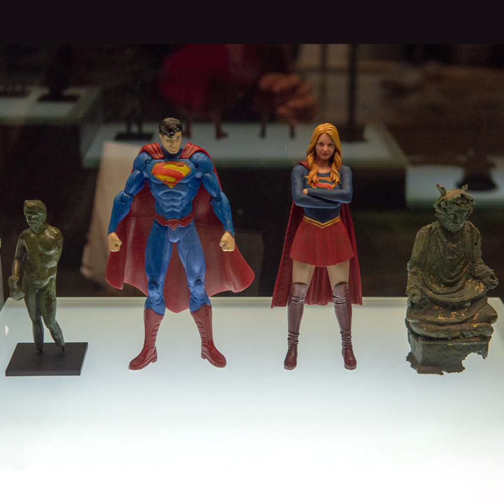 Dans une vitrine une figurine du Superman et de Supergirl sont entourées de deux statuettes antiques en bronze représentant Mercure.