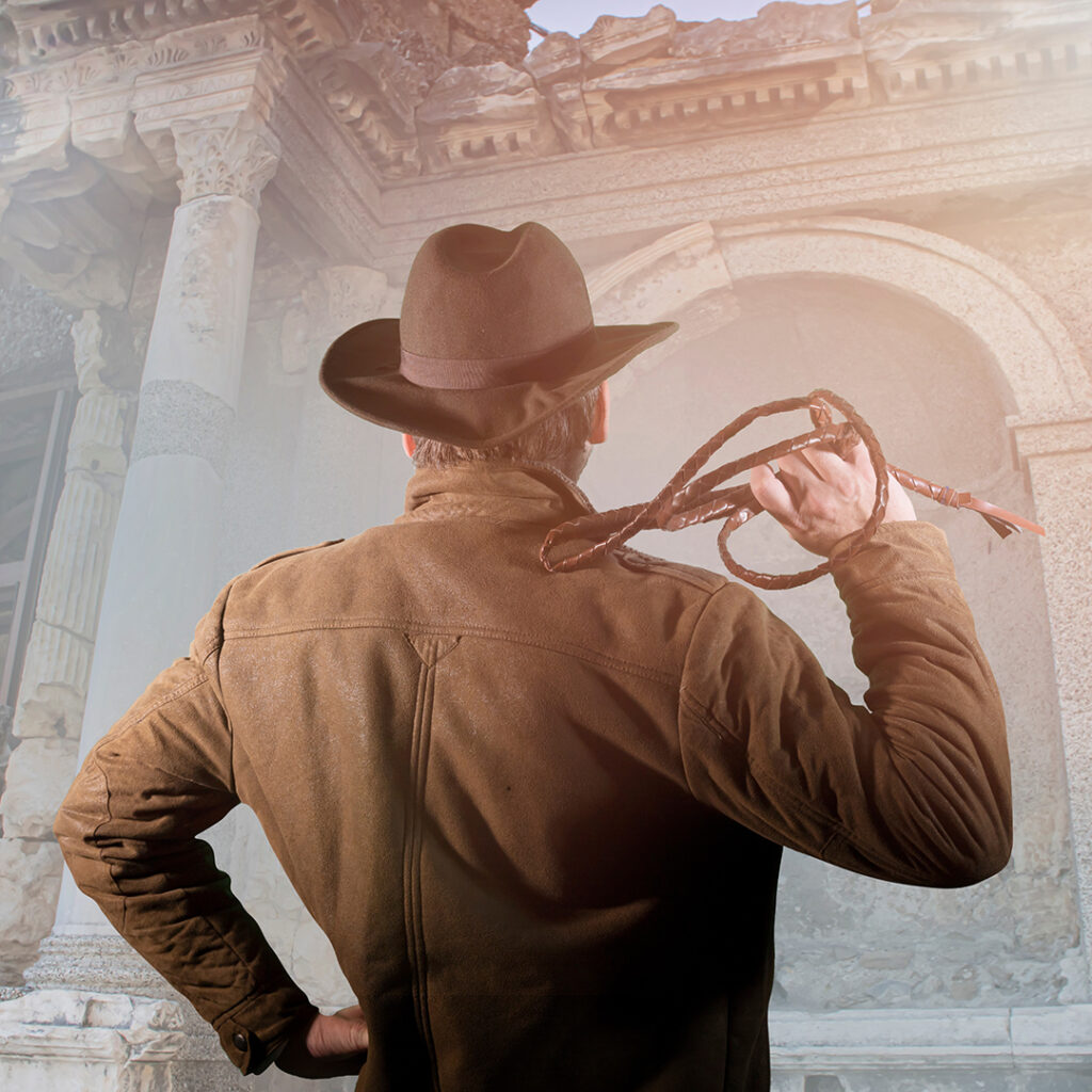 Un homme, vue de dos, vêtu d'un blouson marron, portant un chapeau sur la tête et tenant un fouet dans sa main droite se tient devant des ruines antiques.
