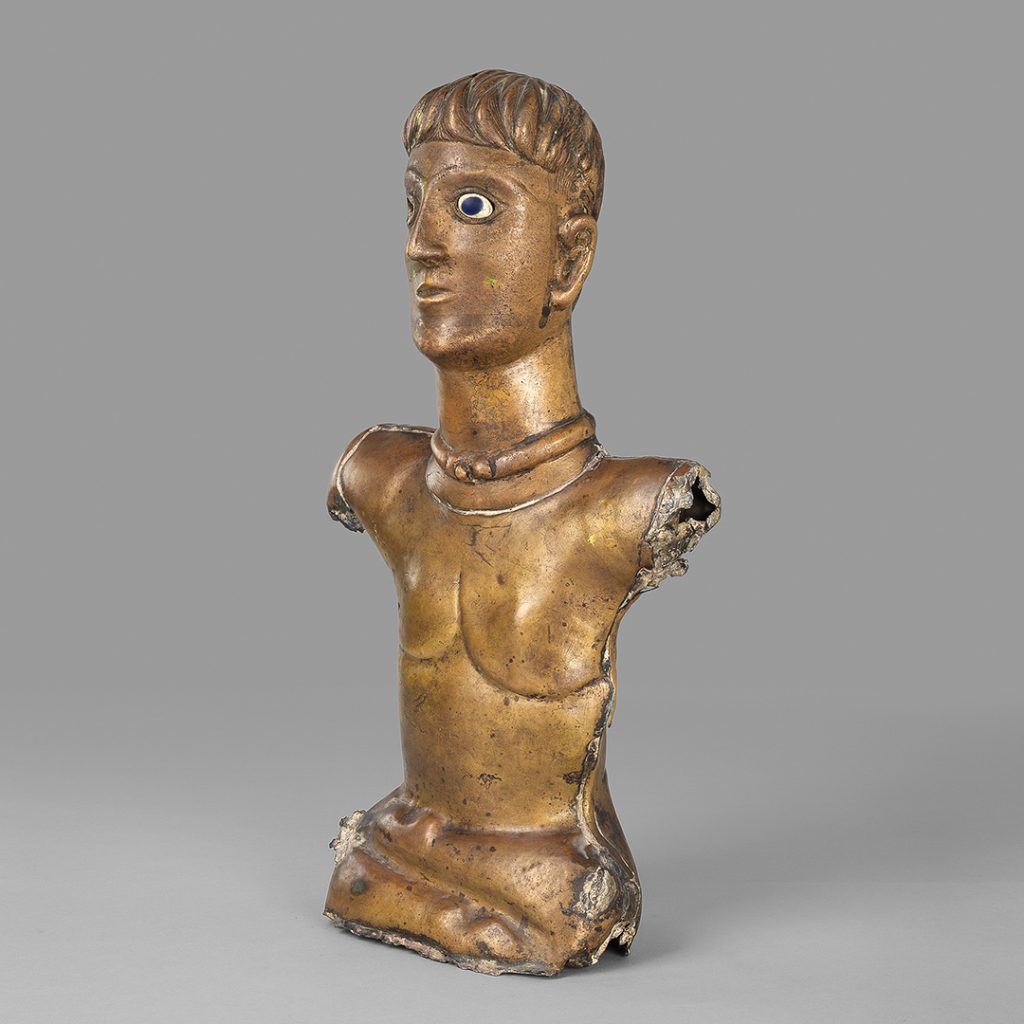 Divinité en bronze assise en tailleur. Elle porte un torque autour de son cou. L'oeil gauche est conservé : il est en pâte de verre blanche et bleu cobalt pour la pupille. Les bras sont manquants. Le torse est nu. Les jambes sont mal proportionnées et les pieds évoquent des pattes de cervidé. 