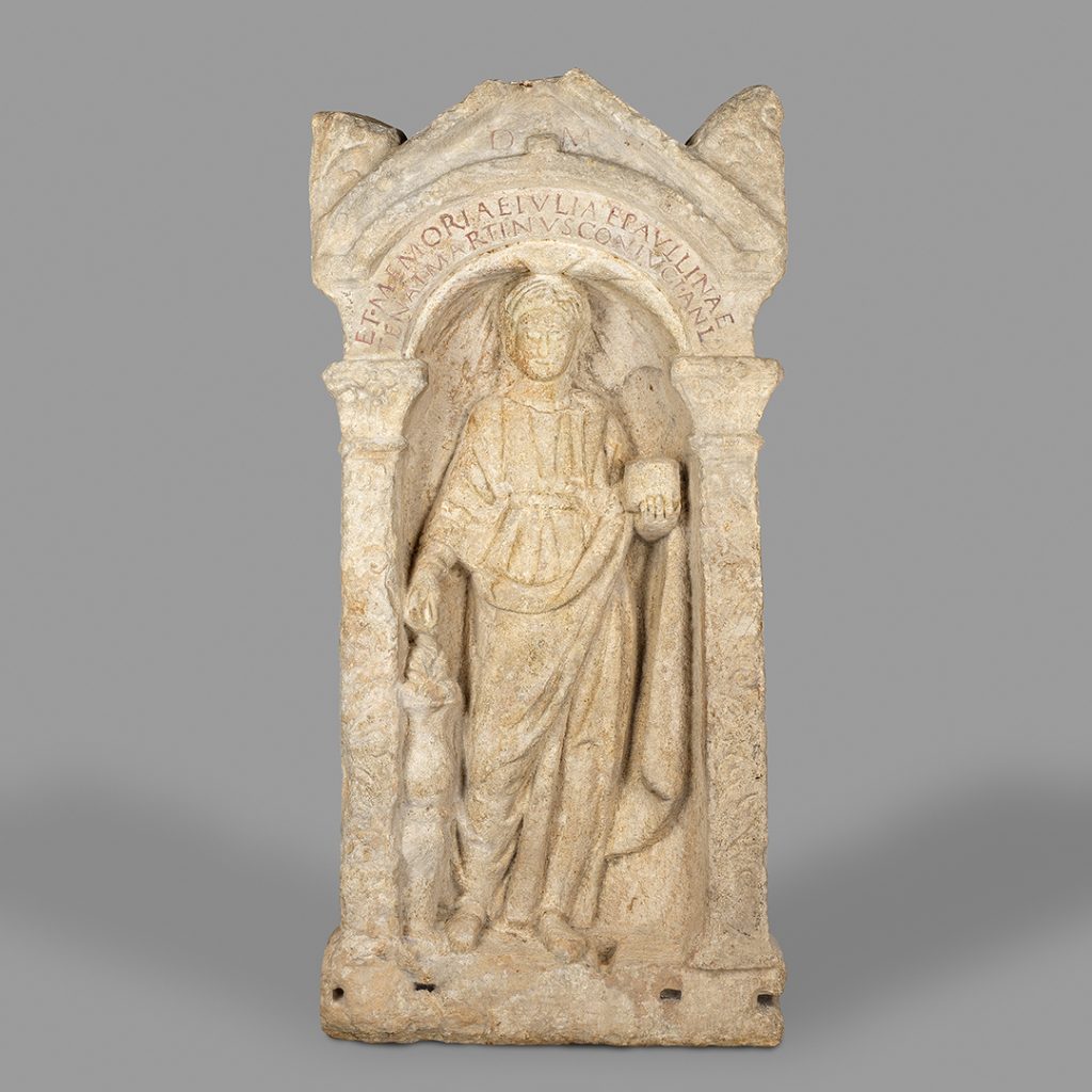 Stèle funéraire montrant, sous une arcade le portrait sculpté en pied de la défunte vêtue d'une tunique et d'un manteau aux larges plis. Elle tient dans sa main gauche une boîte contenant de l'encens qu'elle semble verser sur les flammes d'un autel de la main droite.